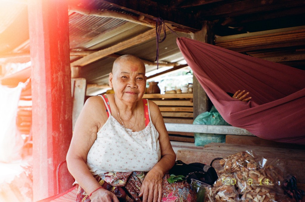 An elderly Cambodian woman with a shaved head sits on a raised dark wood platform, next to a pile of plastic packages filled with Traditional Khmer medicine woody plant materials. She is looking into the camera and smiling slightly. She wears a white sleeveless shirt which is dotted with light red floral patterning, a sarong which is also floral and multicolored in design, a light golden chain around her neck, and a red thread bracelet on her left wrist. In the background is as a red hammock to her right side, where the hand of her granddaughter is poking out; to her left are a series of wooden shelves covered in earthy pots and are covered by a tin roof. 