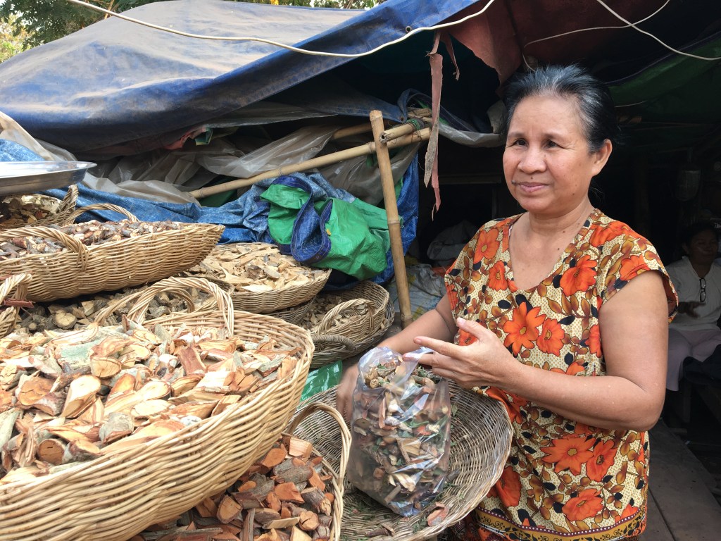A khmer woman with long dark hair tied back behind her neck holds a plastic bag full of Traditional Khmer Medicine woody plant parts over a woven basket. She wears a scooping neck shirt decorated with large orange flowers.  To her left are a cascade of woven baskets full of other Traditional Khmer medicine woody plant parts. In the background, is a shack covered in various blue and green tarps which are layered upon one another as a roof. Faintly on her right side in the distance, there is a man wearing a white shirt, white pants, and glasses sitting on a stool in the shack. 