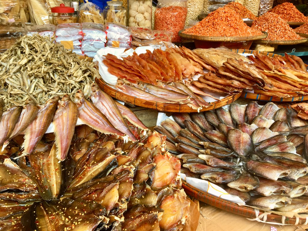 Various large wooden woven plates sit on a merchant table overflowing with traditional Khmer dried fish called trey gneat in a cascading orientation. The dried fish, depending on their size, are either stacked in high piles or meticulously fanned out in a circular pattern to showcase the browns, dark pinks, and oranges of their coloring. Some of the fish are noticeably seasoned with chili flakes. In the background are plastic and glass jars full of other dried foods such as shrimp or other varieties of trey gneat not displayed on the table.

