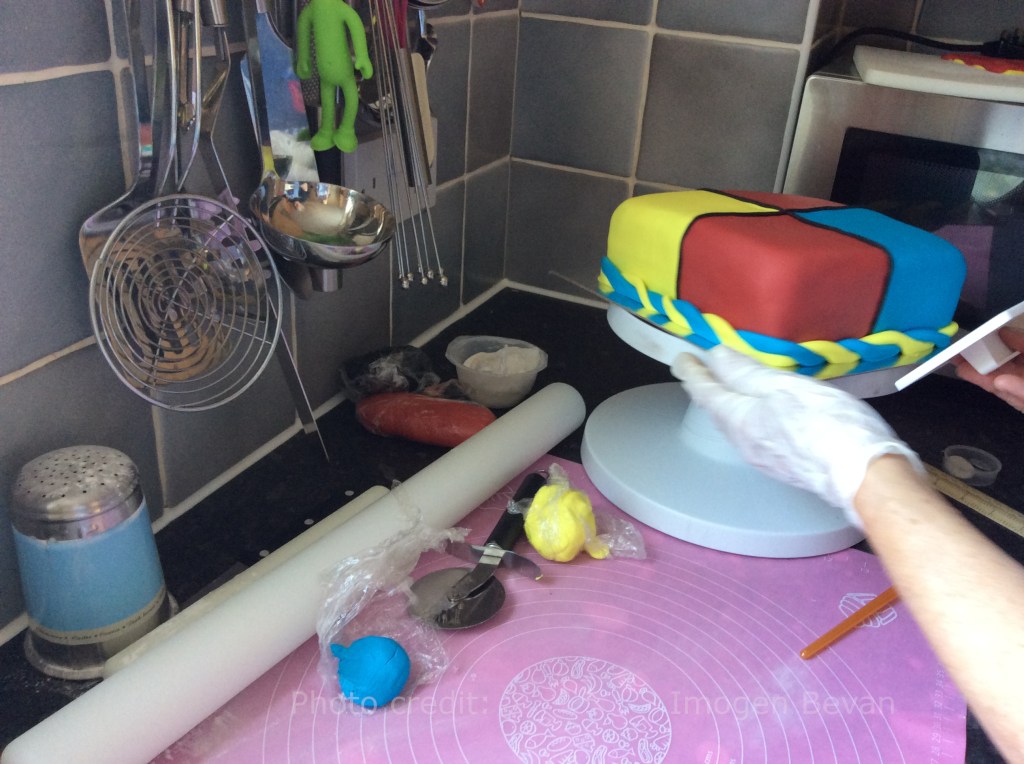 A gloved hand applies red, blue and yellow fondant to a square cake on a cake stand