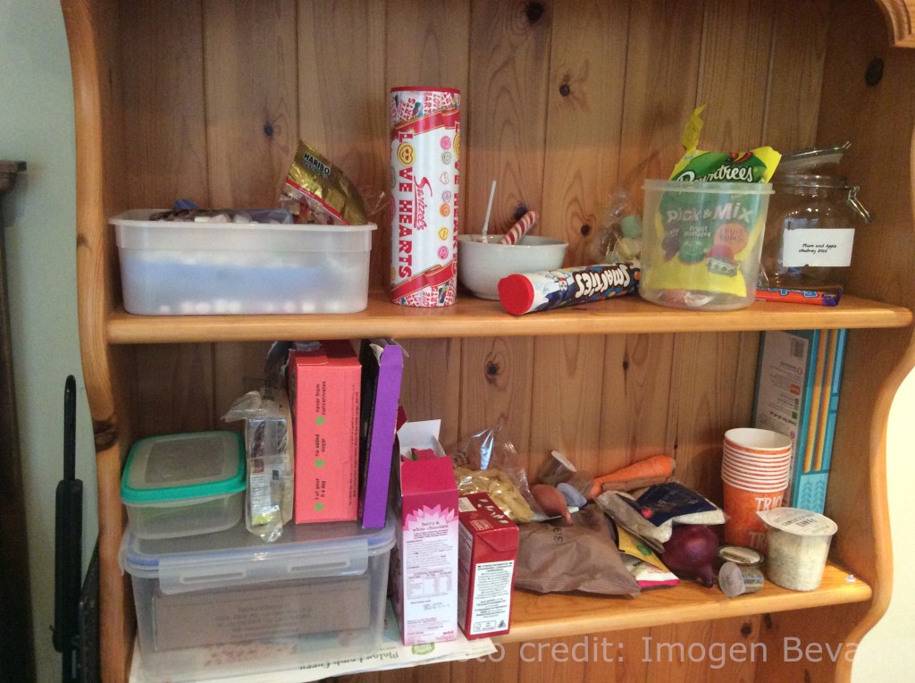 Tubes and bags of sweetse (Love hearts, Smarties and Pick'n'mix) are on the top shelf of a wooden unit